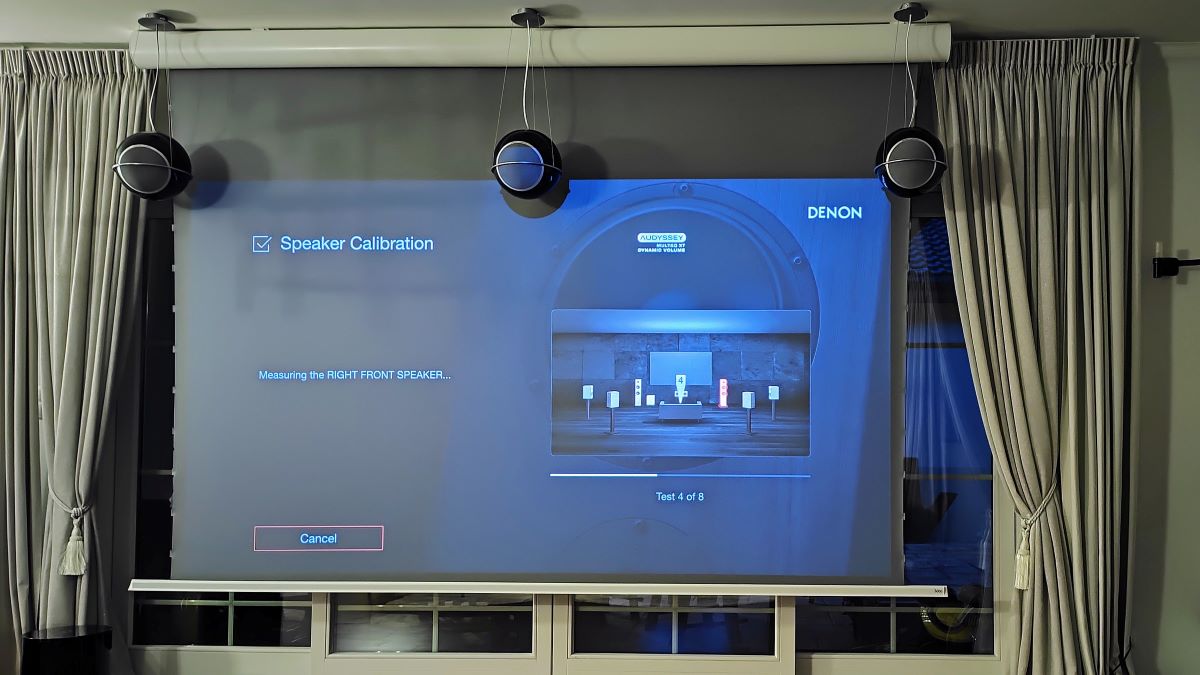 Ekran projekcyjny z napinaczami oraz wiszące głośniki Elipson Planet L w trakcie kalibracji systemu kina domowego